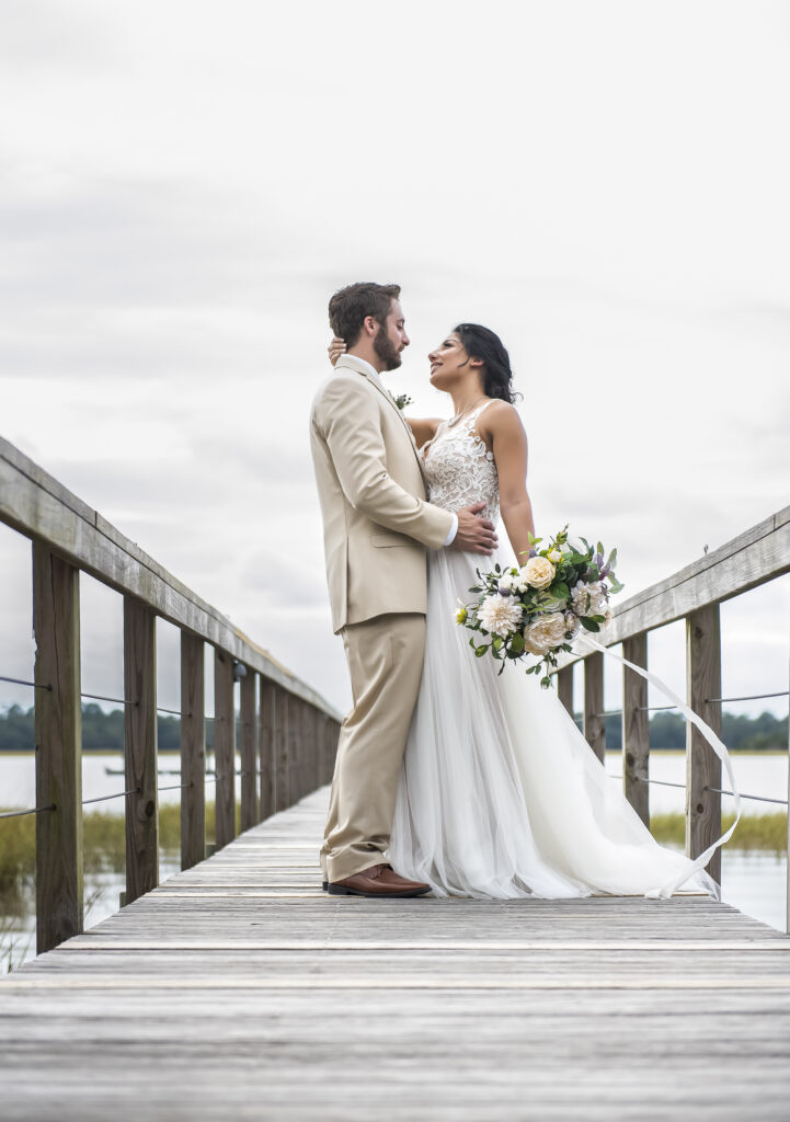 Bride and groom standing on a Charleston dock during their elopement, holding hands and gazing at each other with a soft overcast sky